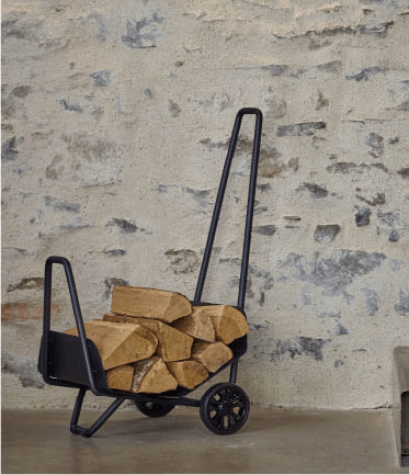 Chariot à bûches en métal noir rempli de bois devant un mur en pierre